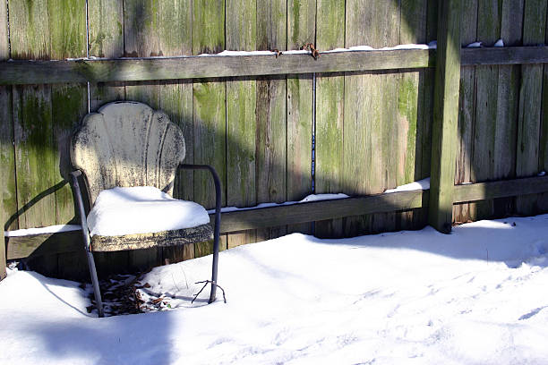 Horizontal shot of a retro chair covered in snow against a wooden fence.