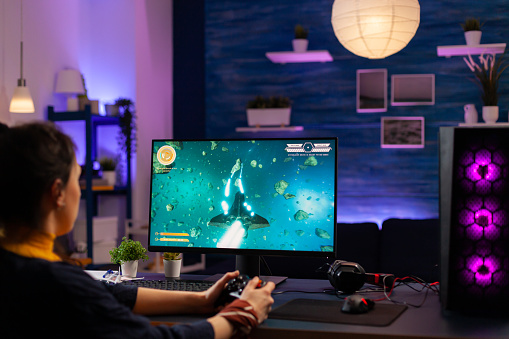 Concentrated player sitting on gaming desk late at night using wireless controller playing space shooter. Virtual streaming cyber performing live tournament using professional equipment