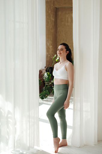 Positive fitness lady with ponytail standing between white curtains in bright spacious room. Athletic female in tight leggings and crop top looking ahead and smiling.