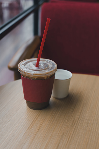 Iced mocha, with ice in a plastic mug, covered with a red paper sleeve, on a wooden table in a certain cafÃ©. Drinking water in a white paper cup