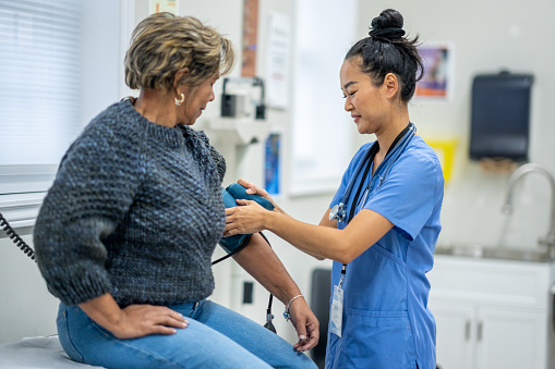 A senior woman of Hispanic decent, sits up on an exam table as her female nurse takes her blood pressure.  The nurse is wearing blue scrubs and making small talk to put the woman at ease.