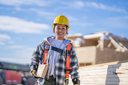 A female construction worker is seen carrying a piece of lumber while working onsite.  She is wearing proper safety equipment and is smiling warmly.