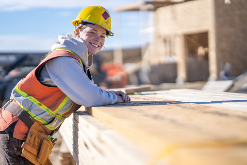 A female construction worker leans on a stack of lumber as she turns to pose for a portrait.  She is wearing proper safety equipment and is smiling warmly.