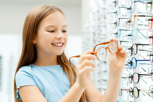 Smiling cute little girl standing in the glasses store while holding eyewear. Health care concept