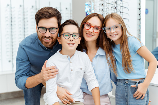 Smiling parents and two kids choosing glasses while posing in optical shop. Health care, family concept