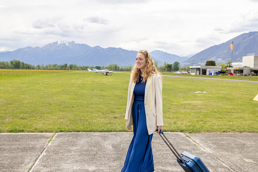 Young corporate businesswoman walking on tarmac toward private jet plane\nShe walks with a suitcase