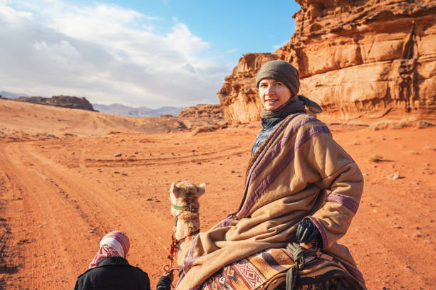 istock Young woman riding camel in Wadi Rum desert, looking back over her shoulder, smiling. It's quite cold so she is wearing traditional Bedouin coat - bisht - and head scarf, mountains far background 1811490536