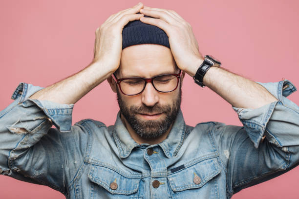 Stressed man in glasses and beanie with hands on head, dressed in a denim jacket, against a pink background.