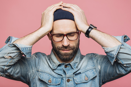 Stressed man in glasses and beanie with hands on head, dressed in a denim jacket, against a pink background.