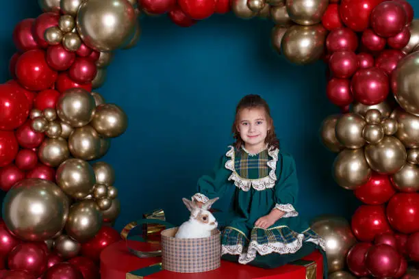 A girl-child in a green dress sits on a huge gift box with a white and brown rabbit against the background of an arch of red and gold inflatable balloons