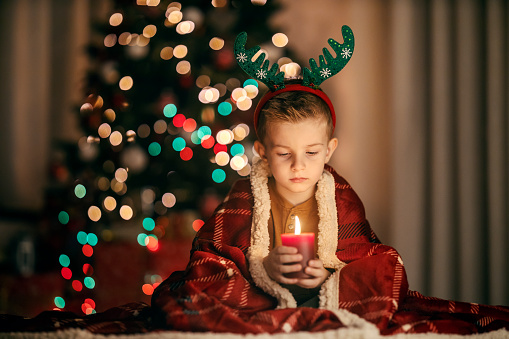 A festive boy is holding a candle on christmas eve at home and looking at it.