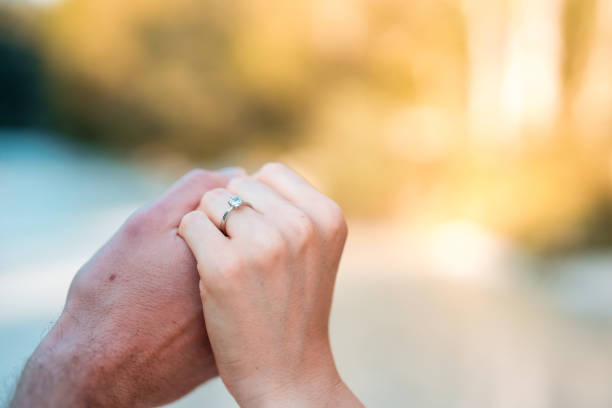 Close-up view of a Caucasian couple holding hands, with a focus on the engagement ring, set against the backdrop of nature.