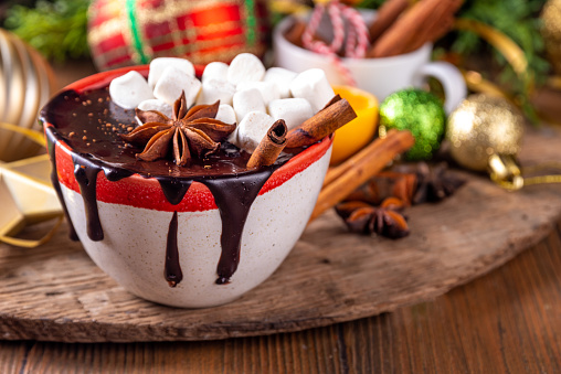 Winter aromatic hot chocolate cup with Christmas background. Mug of dark hot cocoa drink with cinnamon, anise, spices and marshmallow, on cozy Christmas and New Year holiday decorated background
