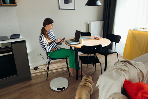 Female with long hair wearing striped shirt and green jeans sitting by the table working on laptop and cleaning her stylish bright apartment with robotic vacuum cleaner in the kitchen, while her cute out is under the table