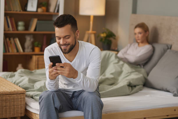 man with smartphone in bedroom, upset woman in the background - infidelity dishonesty heterosexual couple couple imagens e fotografias de stock