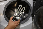 A hand holds a heating element with scale against the background of a broken washing machine. Replacing an old heating element with limescale deposits with a new one on your own.
