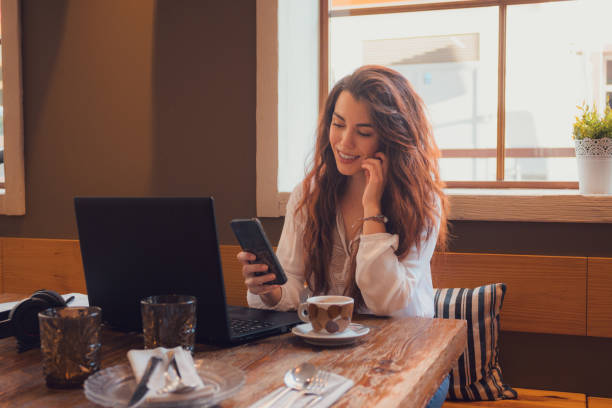 istock Smiling woman looking at her phone while she is working from a restaurant with a laptop 1811659972