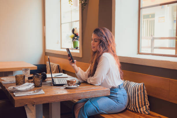 istock Smiling woman looking at her phone while she is working from a restaurant with a laptop 1811659981