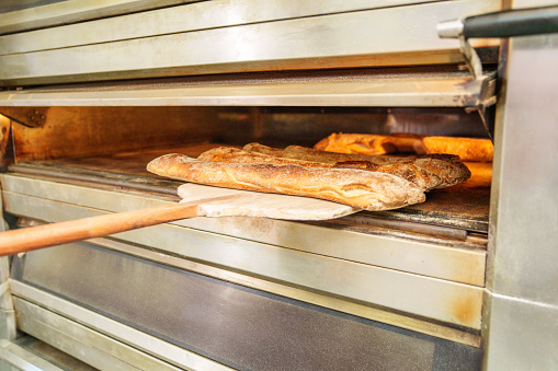 Fresh golden baguette being baked, taken from a hot industrial oven with a wooden baker's peel in a traditional bakery