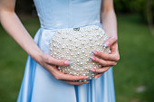 Woman wearing a formal blue satin dress holding a heart-shaped handbag with pearls - detail, close-up view. Selective focus.