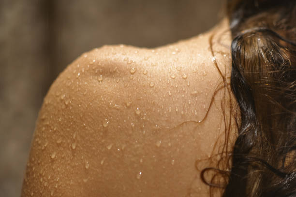 Woman taking shower, with focus on her back and shoulders. Close-up of her skin and water drops. Close-up of a woman taking shower in a bathroom. wet skin stock pictures, royalty-free photos & images