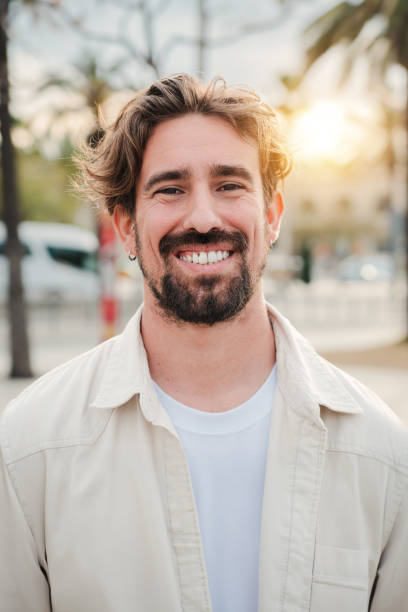 istock Vertical portrait of handsome bearded guy with perfect white teeth smiling and looking at camera standing outdoors. Front view of young adult man with friendly and positive expression. Happy male 1811759932
