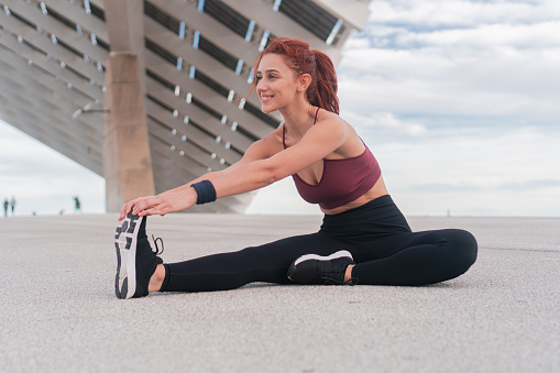 Young woman posing while working out, getting fit and stretching leg sitting and showing a calm attitude and a cheerful smile at the seaside. Sporty female practicing yoga for healthy lifestyle