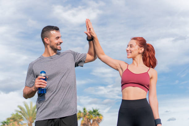 um jovem casal esportivo sorridente e high five para comemorar um treino de sucesso em um dia ensolarado. o esportista está segurando uma garrafa de água. um homem e uma mulher alegres e uma pausa - helathy - fotografias e filmes do acervo