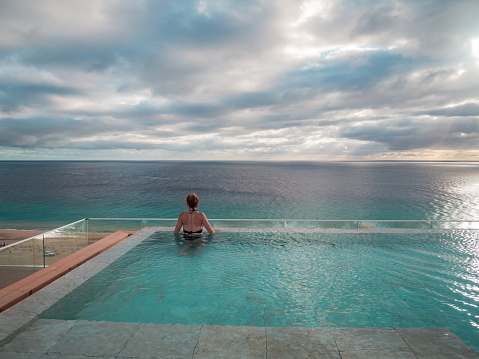 Top rear view of a woman standing in an infinity pool and is looking over the sea - dramatic sky