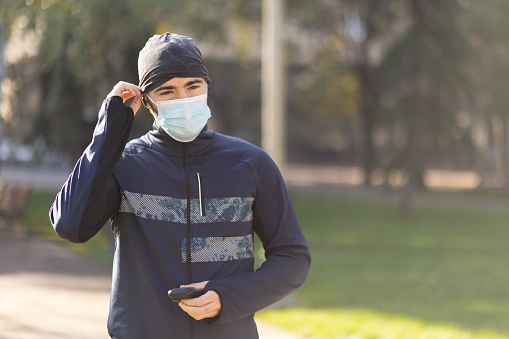 Young male athlete with protective face mask putting on earbuds in autumn park