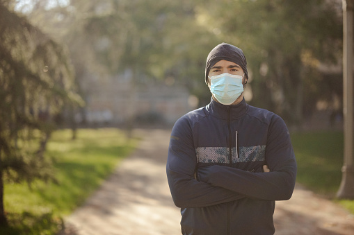 Portrait of young male athlete with protective face mask standing with arms crossed and looking at camera in autumn park