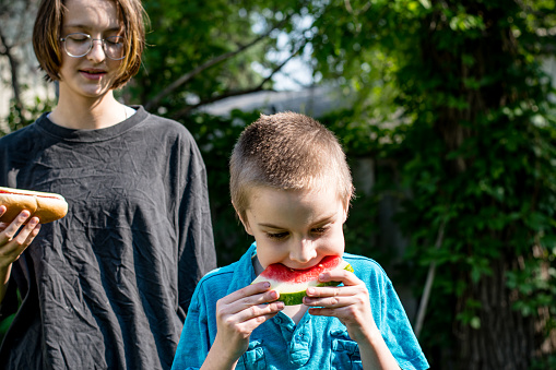 Sibling Connection: Sharing laughter and flavors, a gender non-conforming teen and their younger brother relish a delightful backyard feast, savoring a scrumptious hot dog and refreshing watermelon.