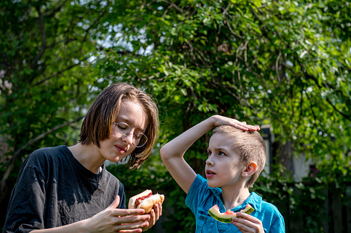 Sibling Connection: Sharing laughter and flavors, a gender non-conforming teen and their younger brother relish a delightful backyard feast, savoring a scrumptious hot dog and refreshing watermelon.