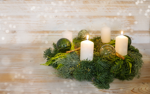 Second advent, natural advent wreath with white candles, two are lit, green Christmas baubles made from glass and cinnamon, wooden background with snowy bokeh, copy space, selected focus