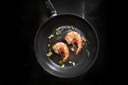 Frying jumbo tiger prawns in olive oil with garlic and herbs in a pan on a black stove top, cooking a gourmet seafood meal with fresh crustaceans, top view from above, copy space, selected focus