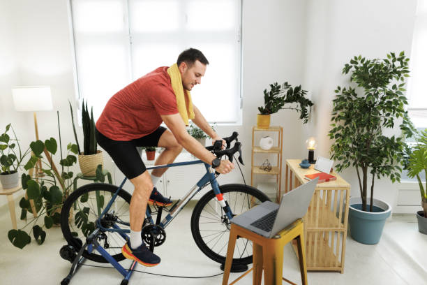 istock Young handsome man exercising on bike at home while using laptop 1812072326