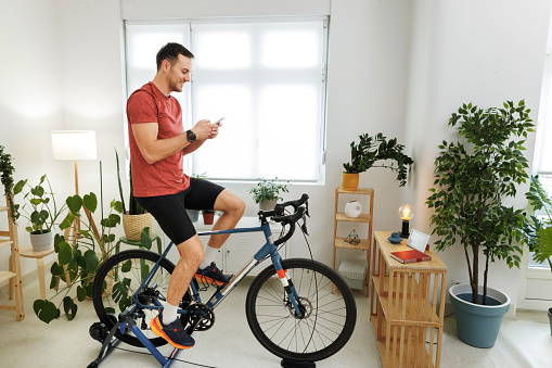 Handsome athletic man using smart phone while riding exercise bicycle at home