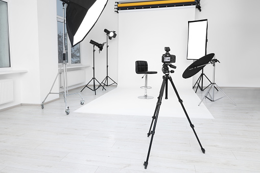 Camera on tripod, bar stool and professional lighting equipment in modern photo studio