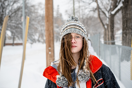 In the quiet charm of a suburban Winnipeg winter, a 13-year-old mixed-race teen exudes sophistication and strength. With a serious expression, she stands as a portrait of serene beauty amidst the cold embrace of the snowy season, donned in casual winter attire and glasses