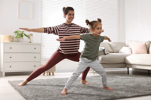 Photo of adult woman wearing black yoga pants and 3 years old baby girl wearing a red t-shirt and white pants doing yoga exercises in living room. They are a ping yoga mat. Shot indoor with a full frame mirrorless camera.