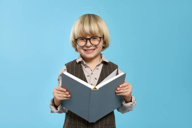 istock Cute little boy in glasses reading book on light blue background 1812164570
