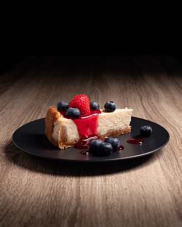 Classic New York Cheesecake dressed with a strawberry, strawberry topping and blueberry on a black plate on a wooden background in studio lighting