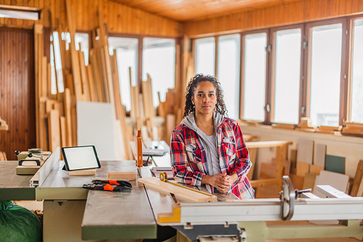 A young female dark-skinned carpenter looking relaxed at a carpentry. She is looking to the camera, dressed casually and has a ponytail, while leaning on a table full with materials and equipment to work with wood.