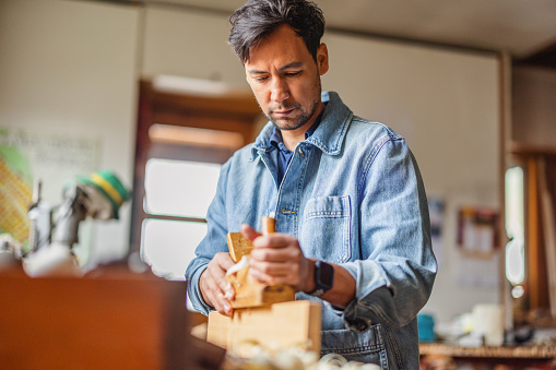 A focused Latin male carpenter dressed casually with a jeans jacket uses a block plane over a rectangular piece of wood to remove any imperfections off the wood.