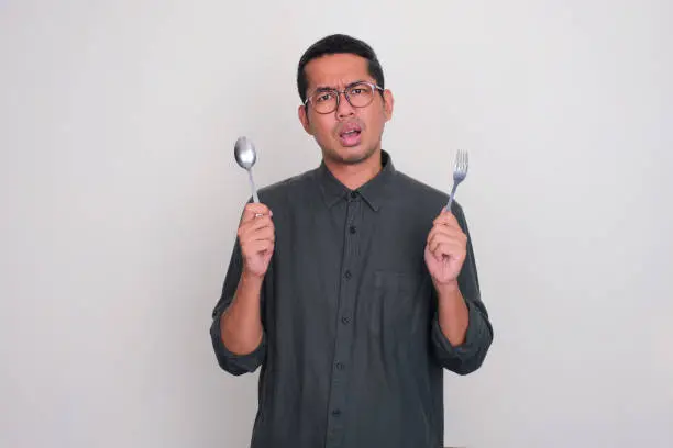 Adult Asian man showing confuse expression while holding spoon and fork