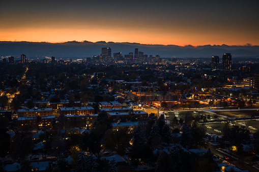 Aerial cityscape of Denver, Colorado at nightfall on a snowy evening, looking across houses and apartment buildings towards the downtown skyline with the Rocky Mountains in the distance.