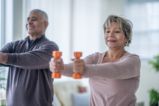 A husband and wife of Hispanic decent, stand in the comfort of their own home with dumbbells in hand, as they work out together.  They are both dressed comfortably and are smiling as they work to stay fit and healthy.