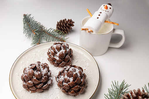 Christmas tree cones haped cookies with chocolate flakes with powdered sugar, a delightful homemade dessert for the New Year. A cake in the shape of a pine cone and a marshmallow snowman in a cappucci