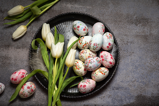 Beautiful white tulips with colorful decoupage eggs in bowl on light gray stone background. Spring and Easter holiday concept with copy space