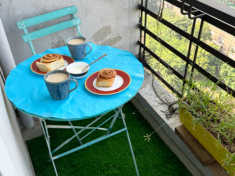 Stock photo showing an apartment balcony decorated with potted plants and artificial green grass turf. A metal circular patio has been set for  breakfast with plates containing cinnamon buns and mugs of milky coffee.
